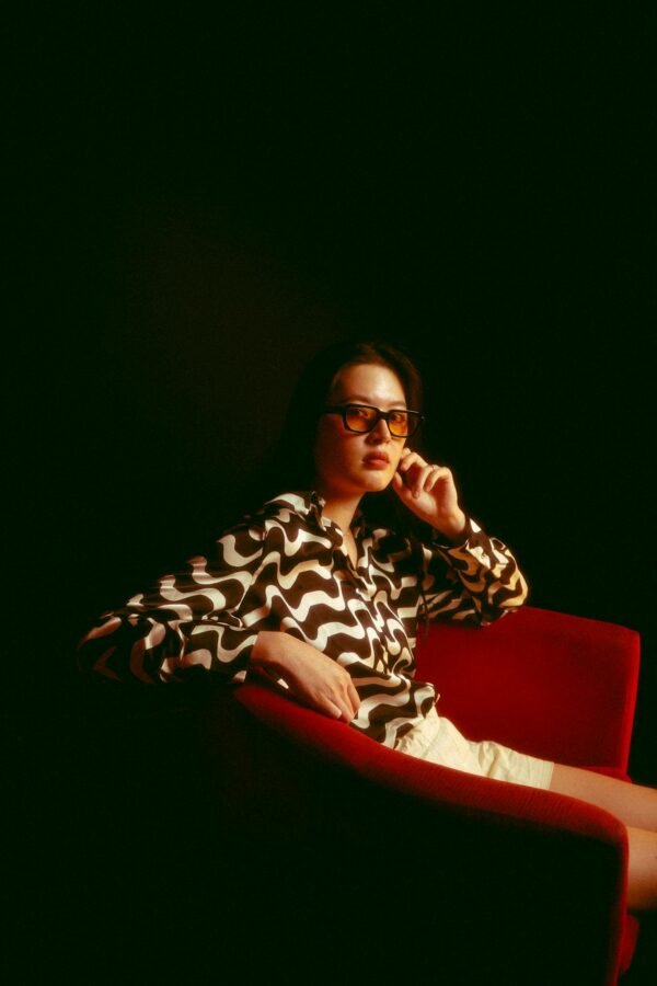 A fashionable woman wearing sunglasses sits on a red armchair in a studio setting.