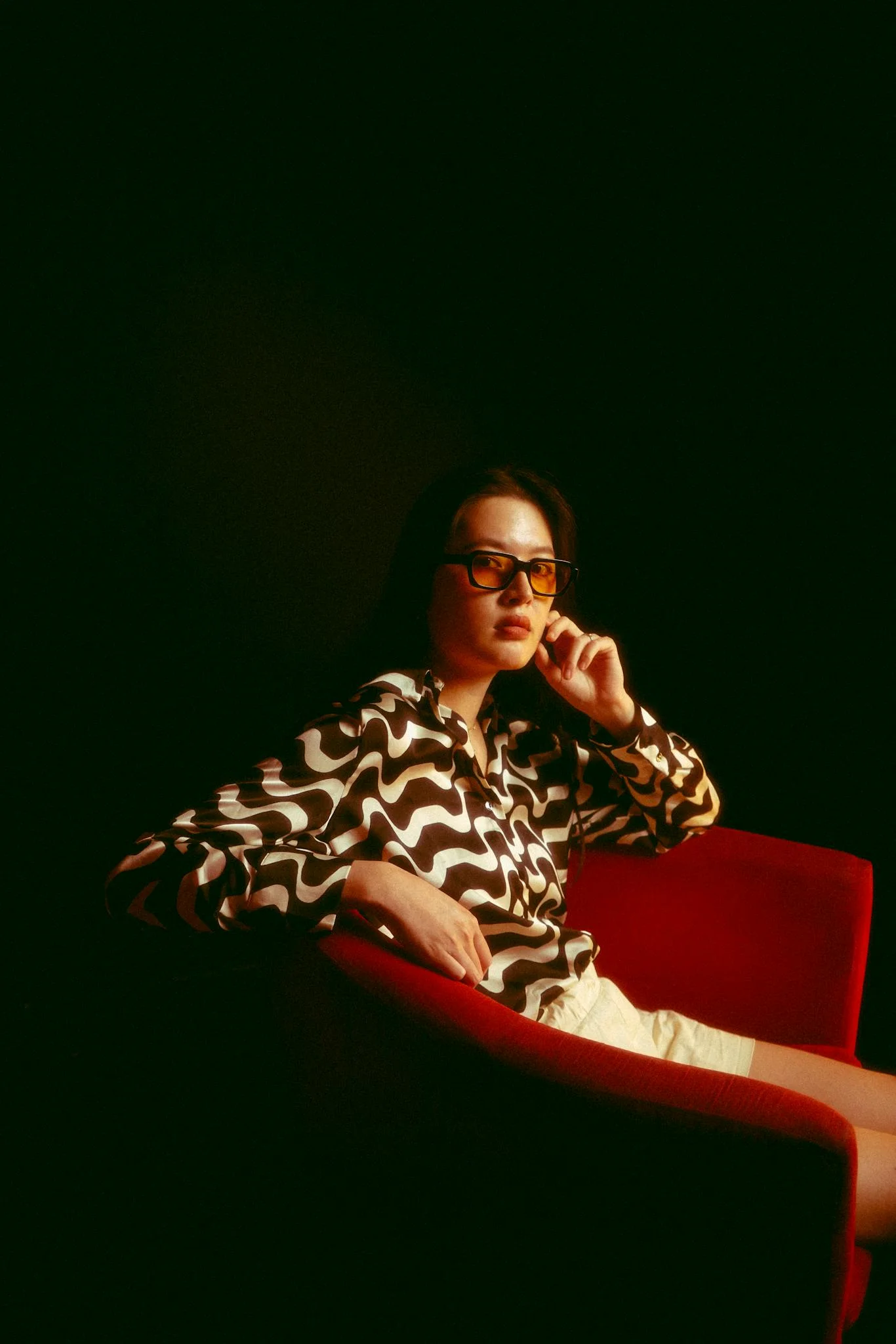A fashionable woman wearing sunglasses sits on a red armchair in a studio setting.