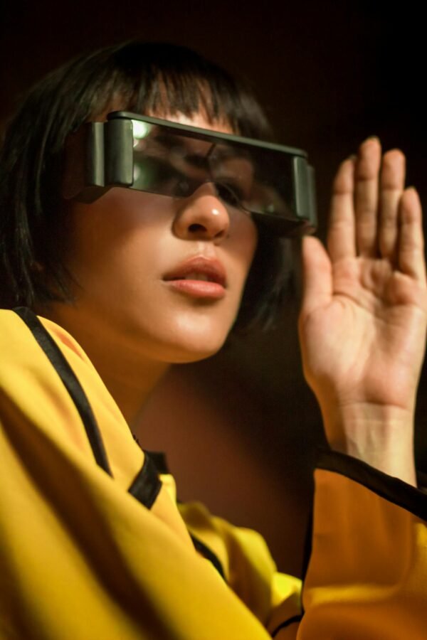 Woman wearing futuristic sunglasses and a yellow outfit, posing indoors with a trendy vibe.
